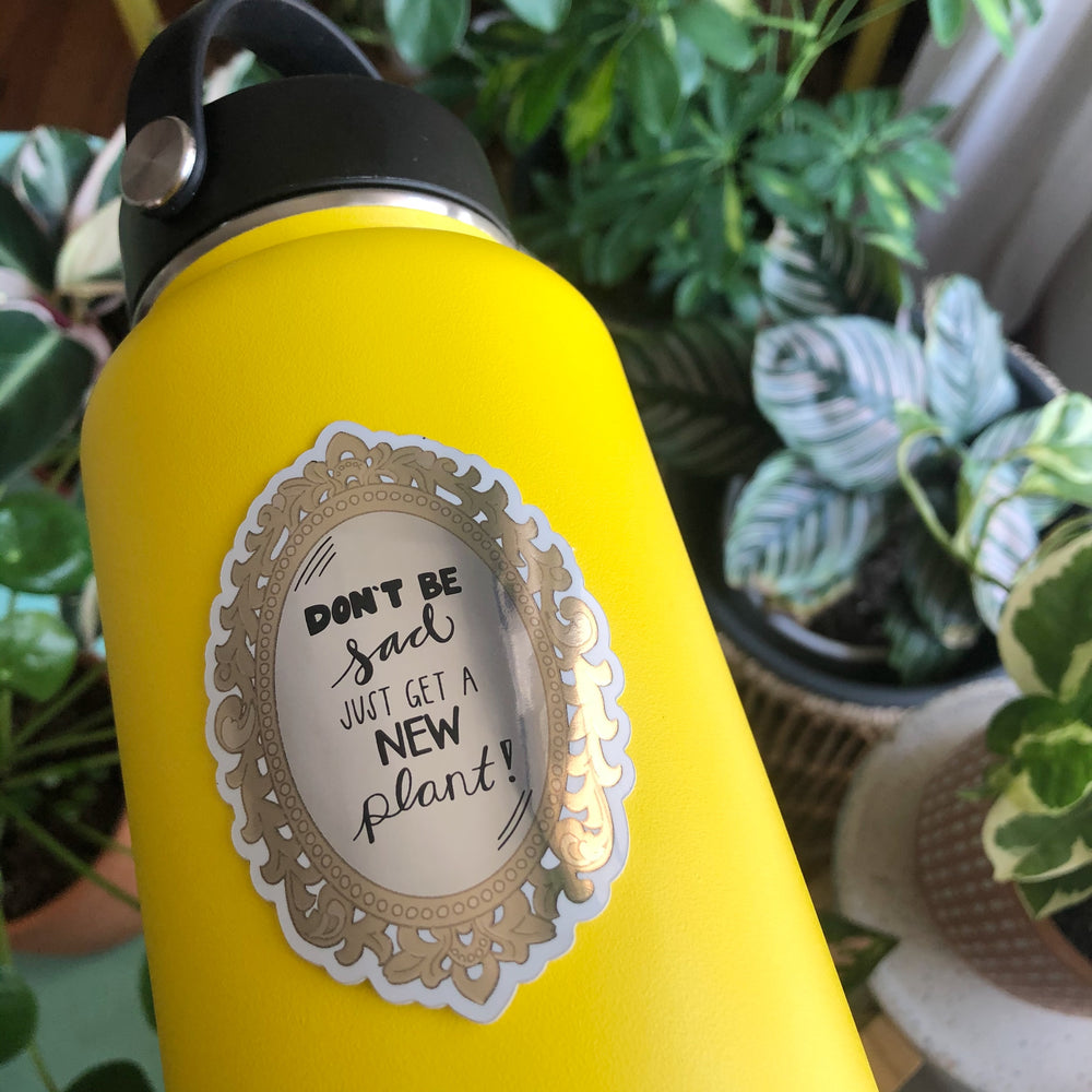 Decorative sticker with text 'Don't be sad, just get a new plant!' stuck on a yellow water battle with green plants in the background