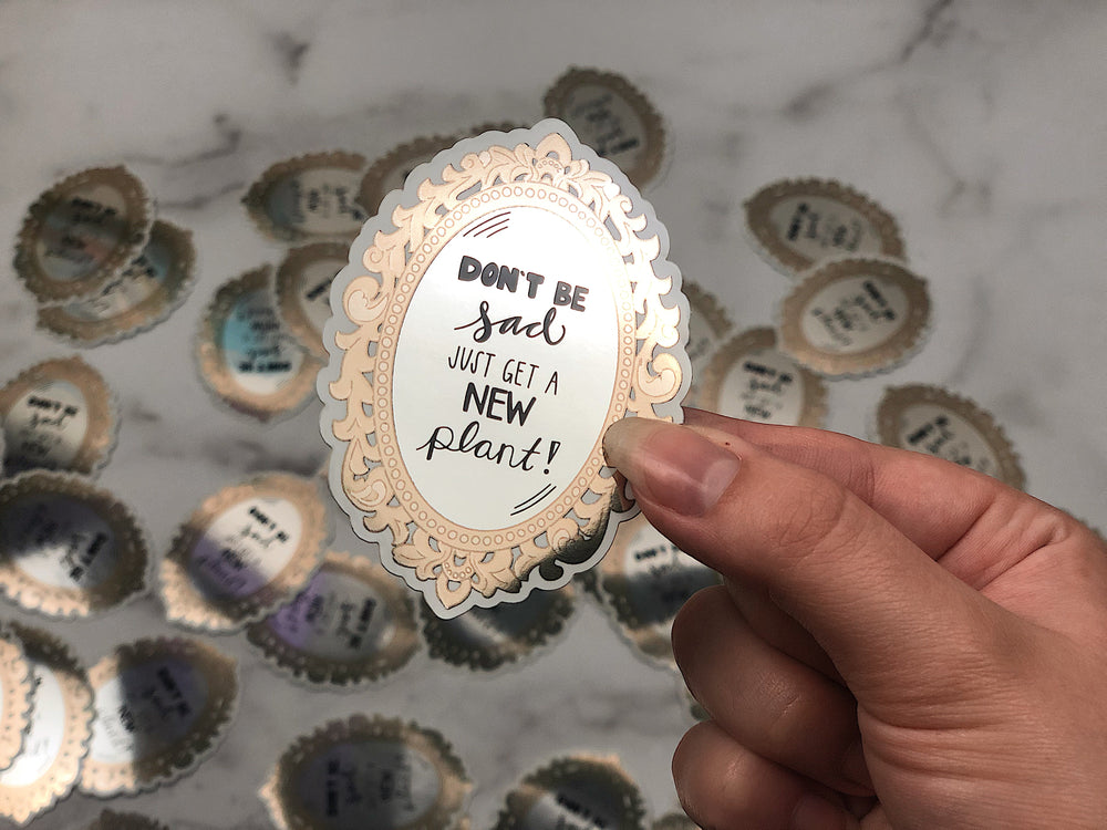 Decorative sticker with text 'Don't be sad, just get a new plant!' held between someones fingers