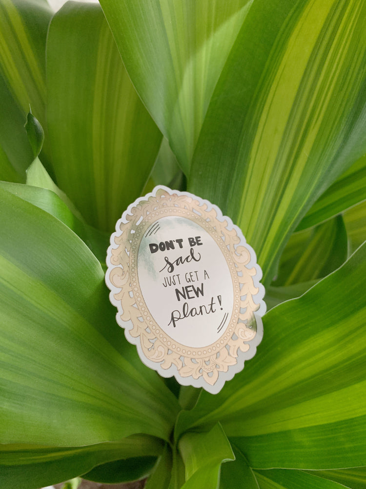 Decorative sticker with text 'Don't be sad, just get a new plant!' held in front of green leaf