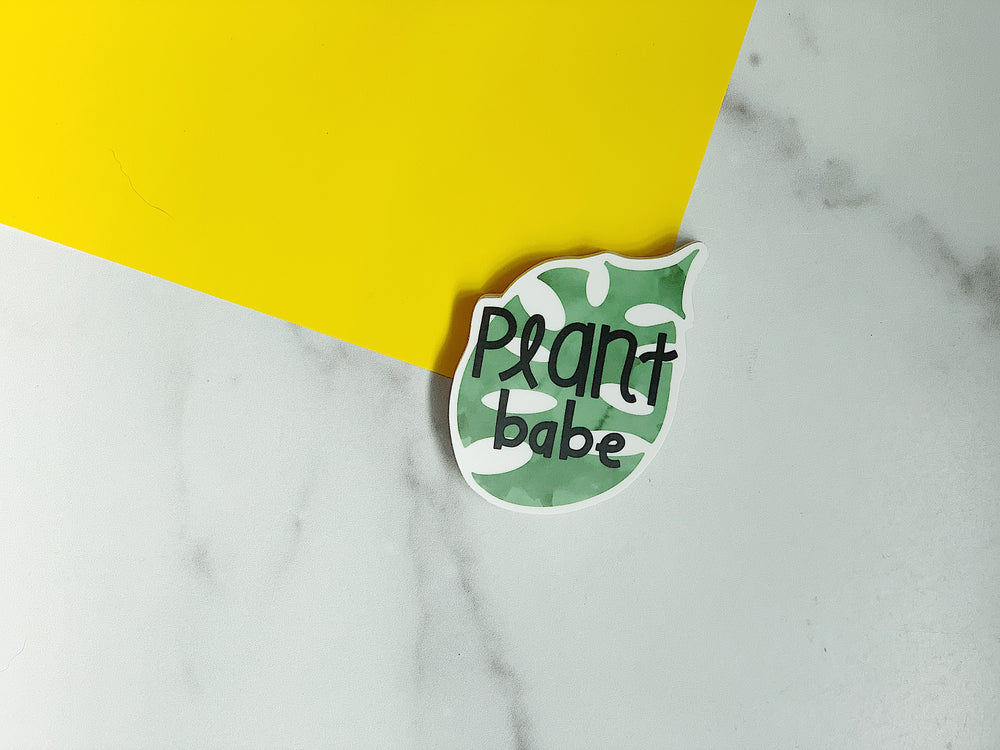 Sticker with 'Plant babe' text on a green leaf design against a yellow and marble background