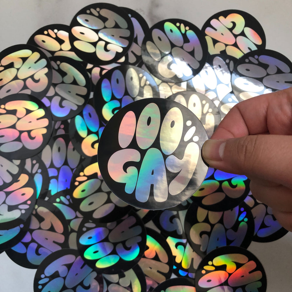Decorative sticker with text '100% Gay' held in front of a pile of the same sticker design