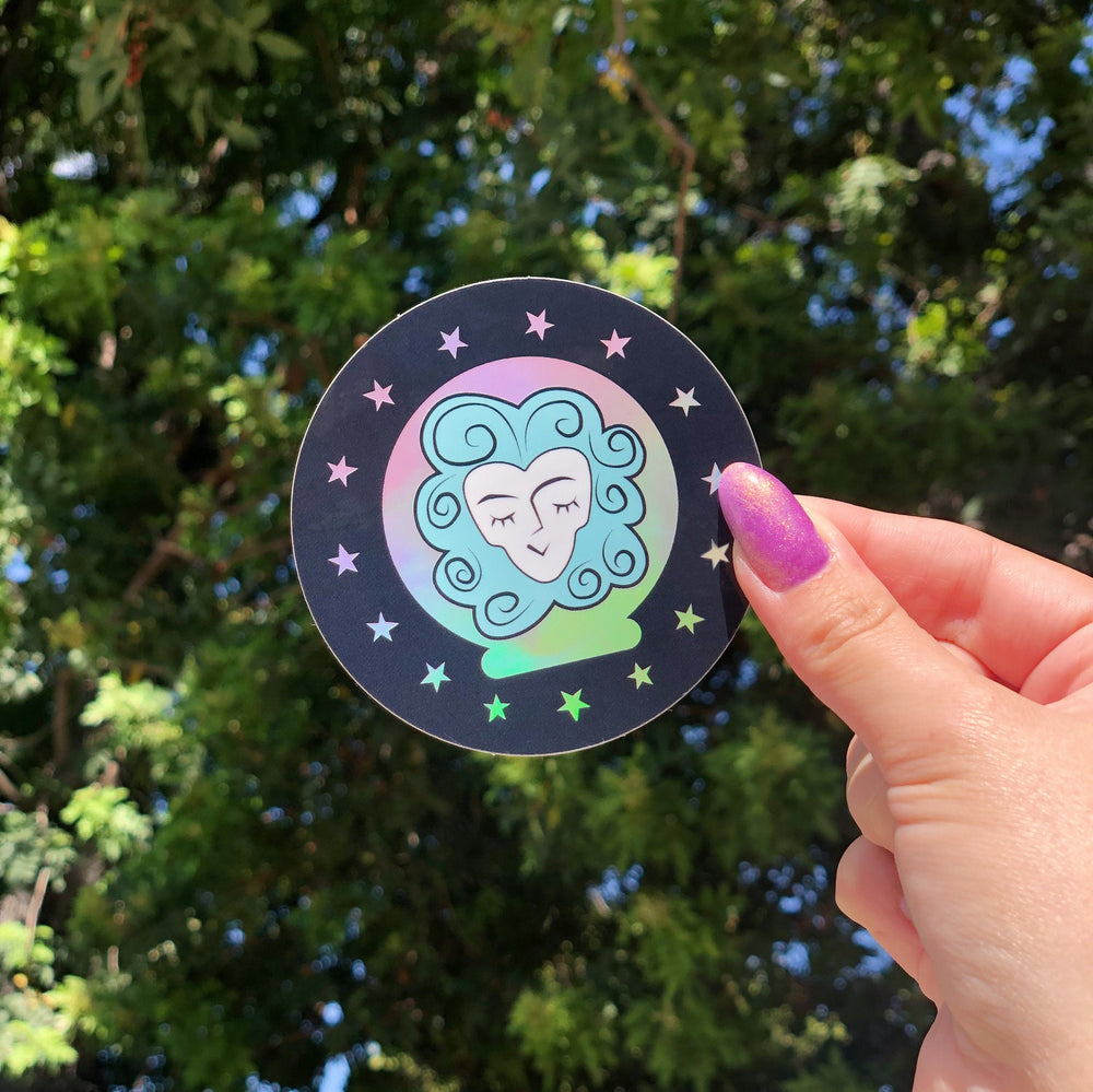 Madame Leota Sticker held with trees in the background. The Sticker is holographic in some parts which look like beautiful rainbow colors.