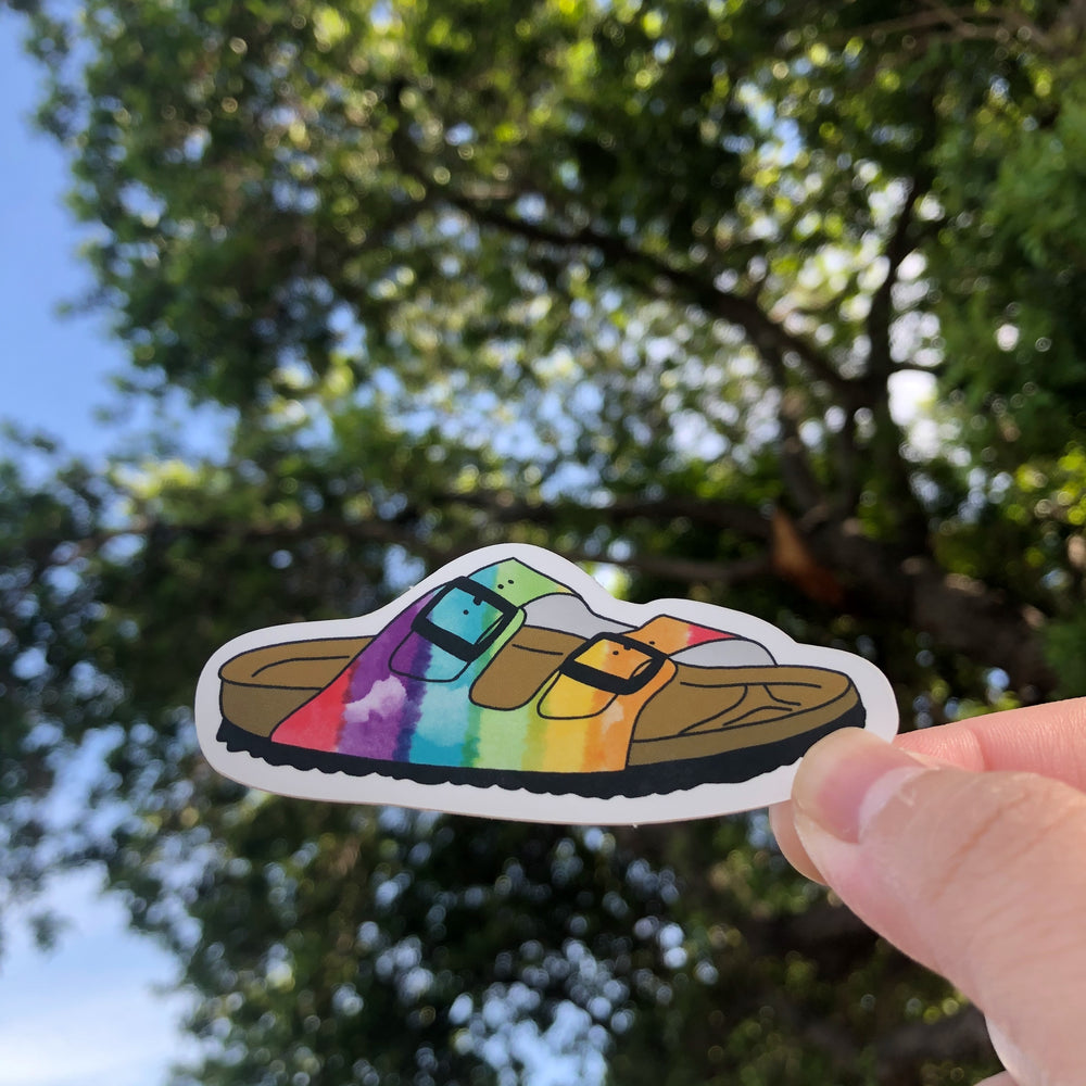 RainbowSandal Sticker being held with a tree and sky in the background