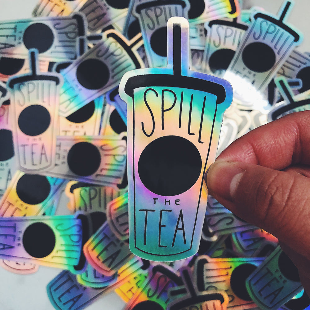Sticker in the shape of a cup with lid and straw that says 'Spill The Tea" held in front of a pile of the same sticker